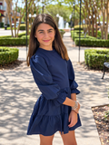 A young girl with long brown hair stands on a paved pathway in a landscaped park area, wearing a navy blue mini dress with three-quarter ruffled sleeves and a tiered skirt.